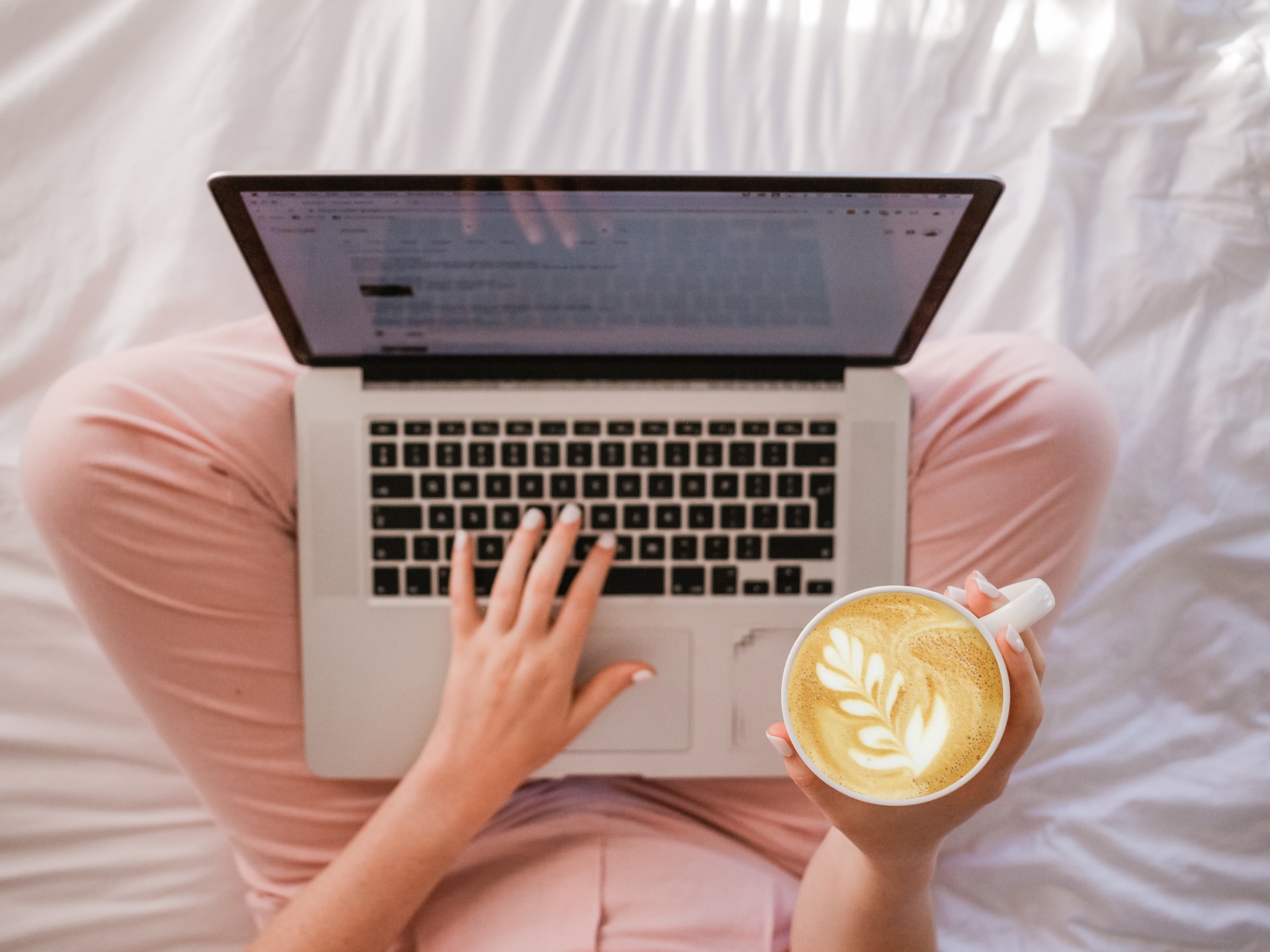 Person sat using a laptop on their bed with a coffee in one hand